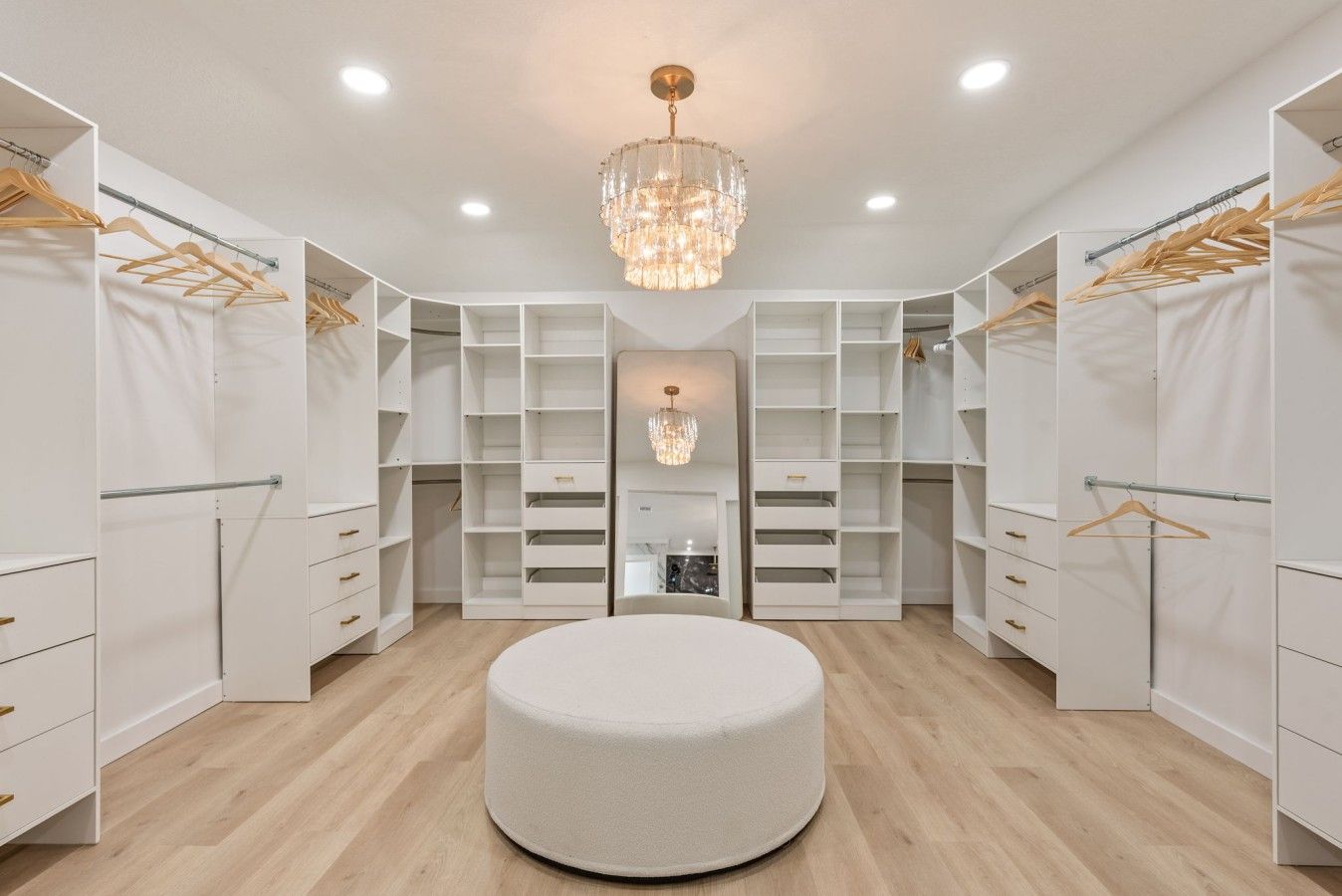 A walk-in closet with white shelving, wooden hangers, and a central round ottoman.