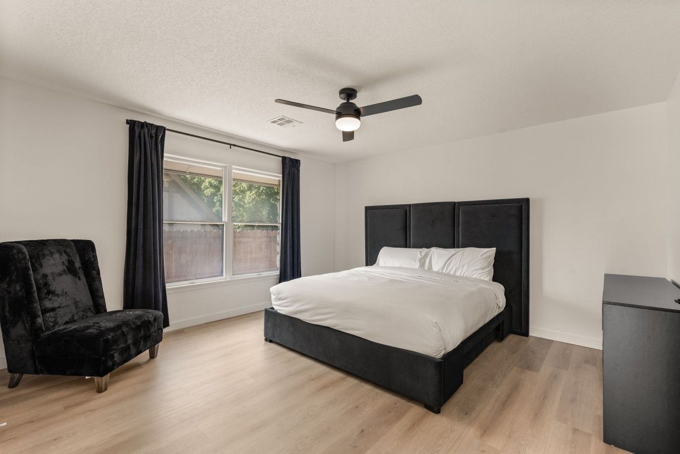 Un dormitorio con cama, una silla, un escritorio, un ventilador de techo y una ventana con cortinas.