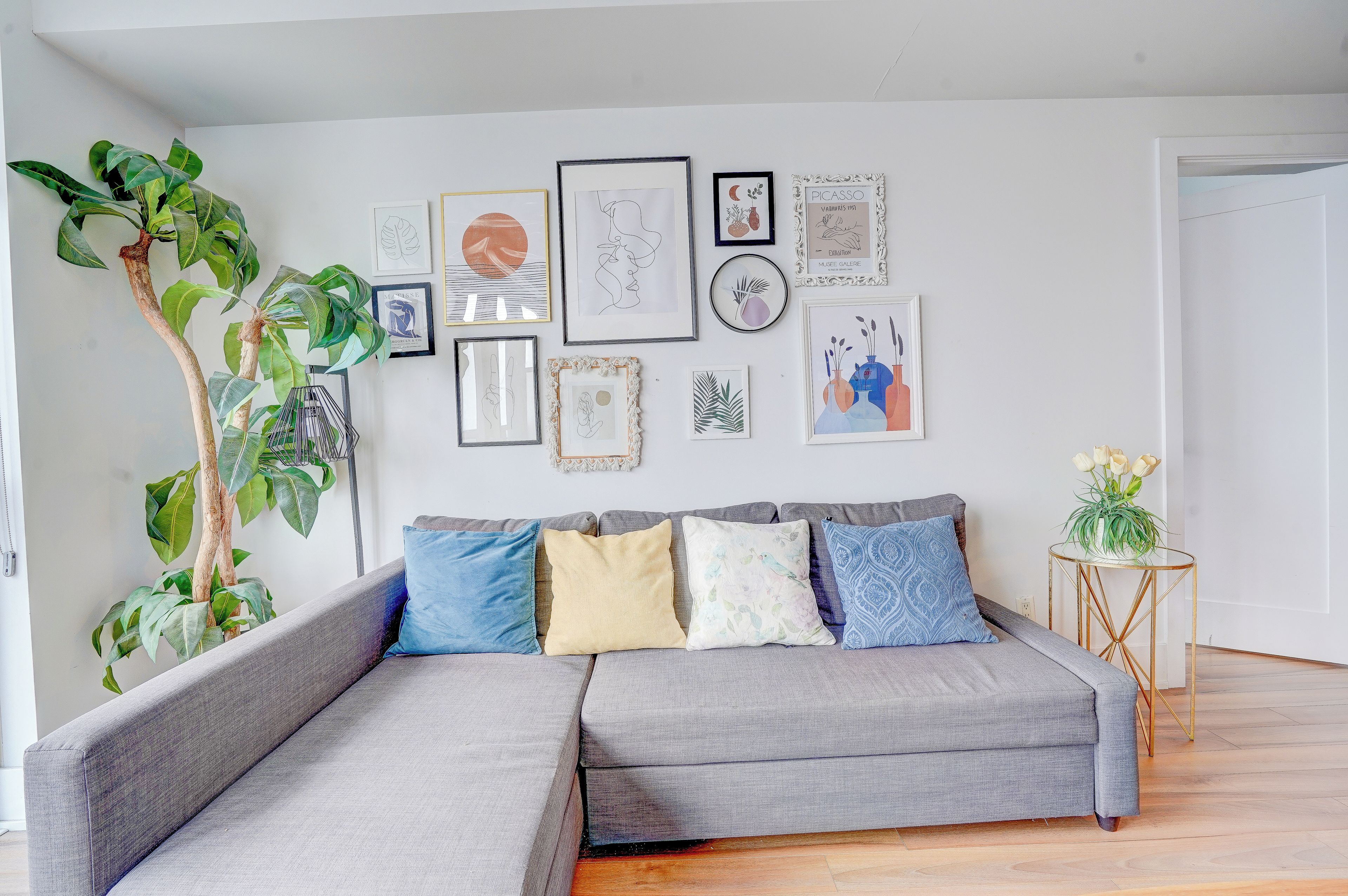 A modern living room with a grey sectional sofa, a large green plant, and a variety of framed artworks on the wall.