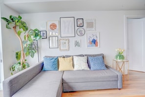 Una sala de estar moderna con un sofá seccional gris, una planta verde grande y una variedad de obras de arte enmarcadas en la pared.