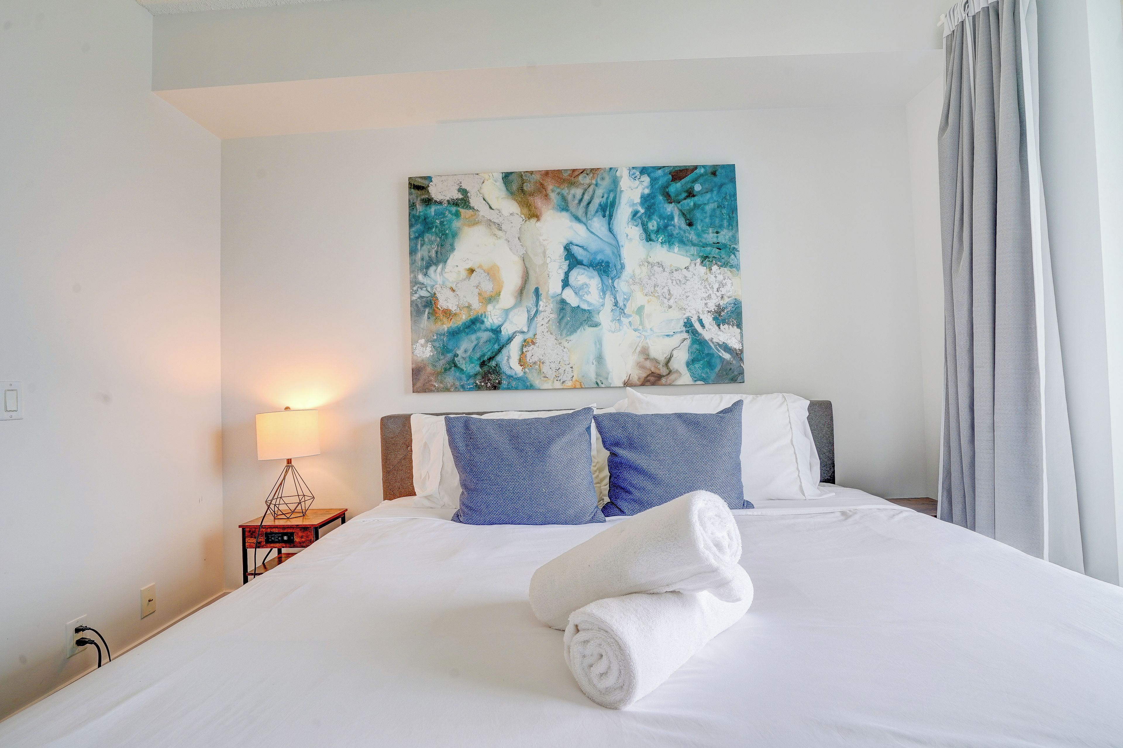 A hotel room with a bed, a painting, a nightstand, and a lamp.