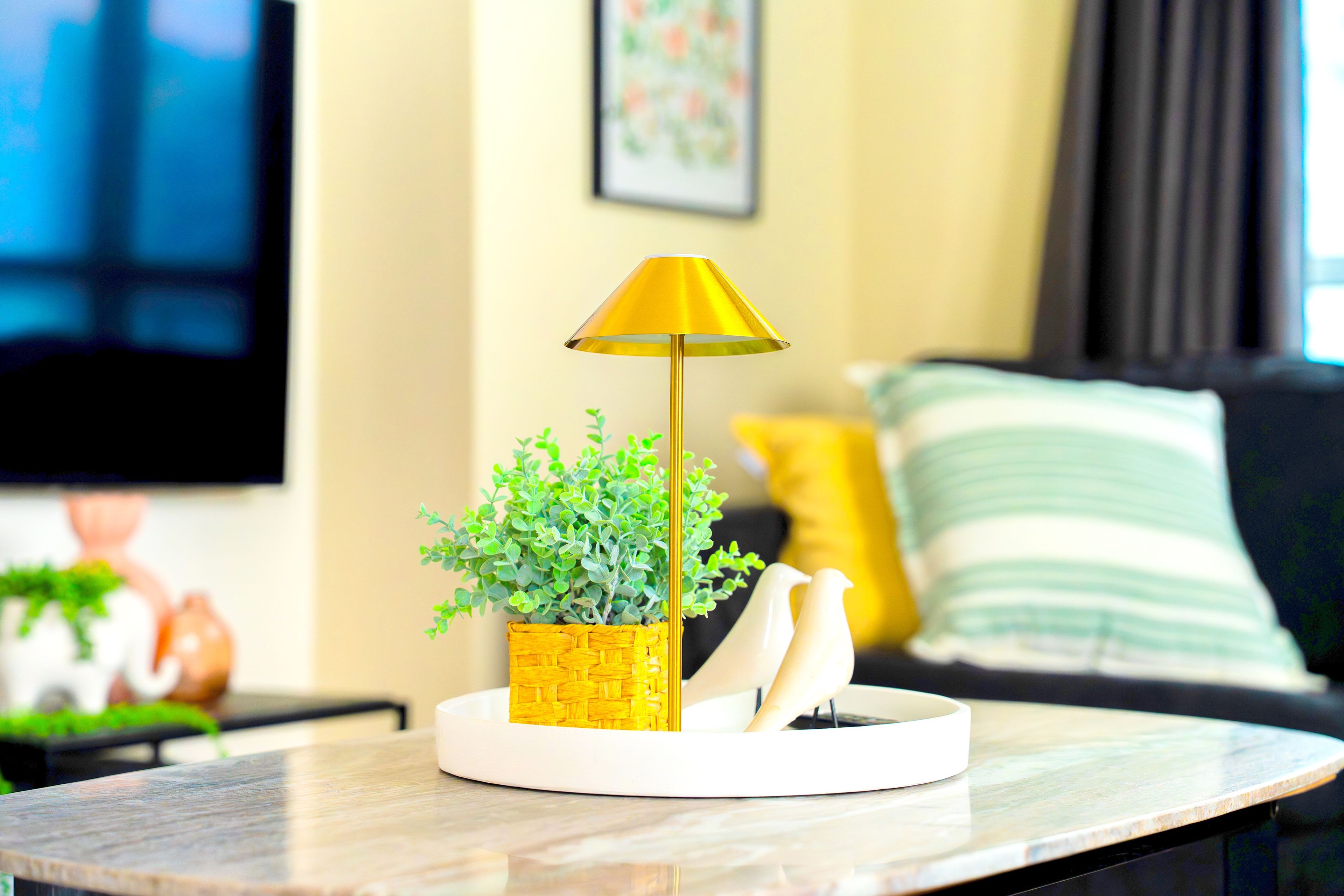 Una mesa con una lámpara, una planta en maceta y dos figuritas blancas.
