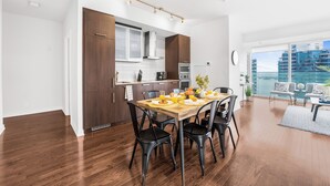 A modern kitchen with dark wood cabinets, a dining area with a wooden table and black chairs, and a view of a cityscape through large windows.