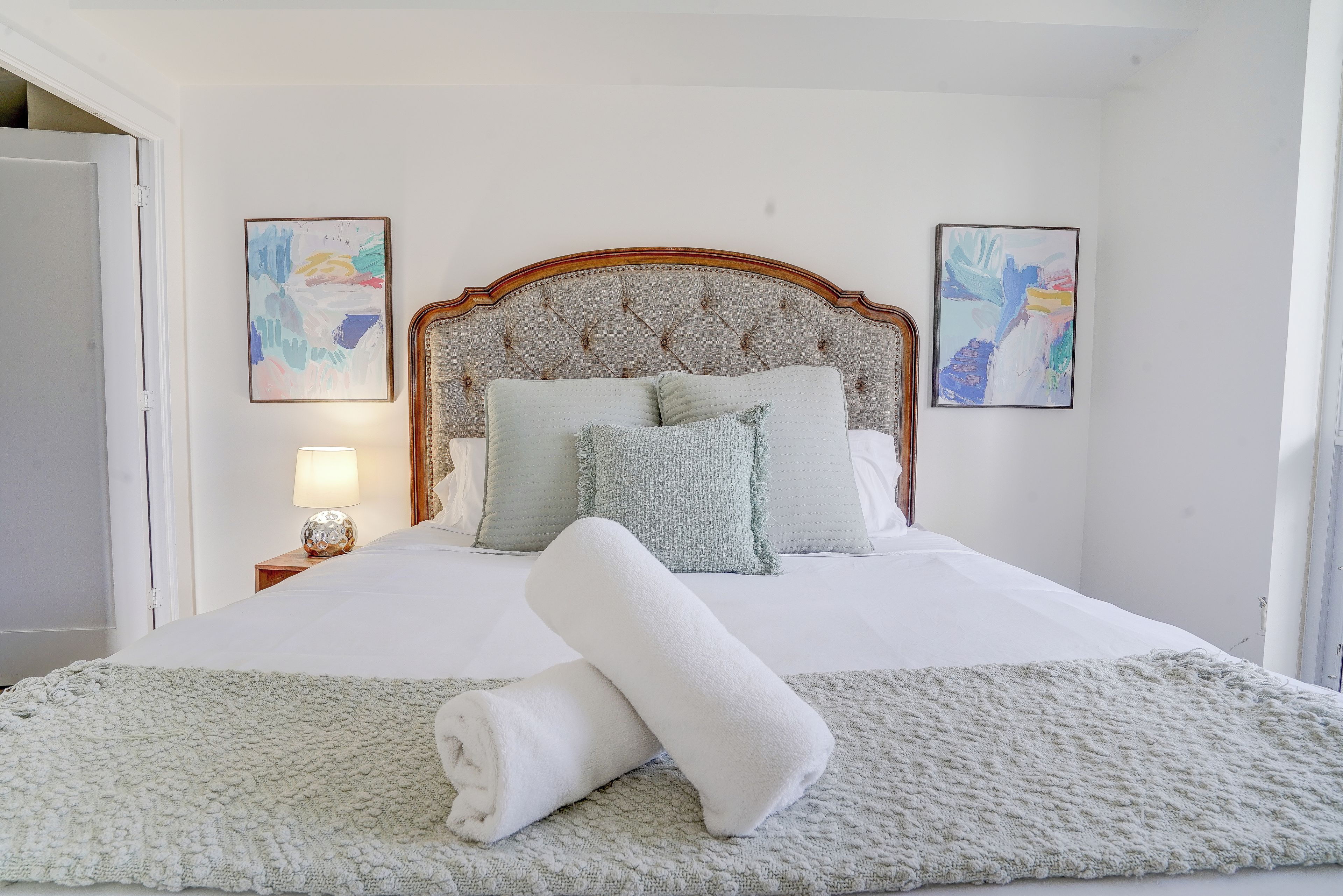 Una cama bien hecha con una colcha blanca, almohadas y una toalla blanca enrollada. Hay dos cuadros enmarcados en las paredes.