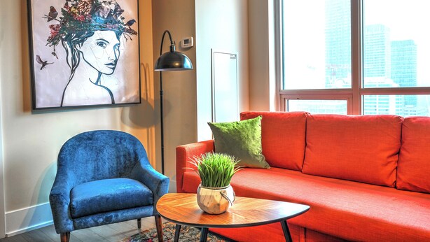 Un salon avec un fauteuil bleu, un canapé rouge, une petite table sur laquelle est posée une plante, un lampadaire et une grande fenêtre donnant sur la ville.