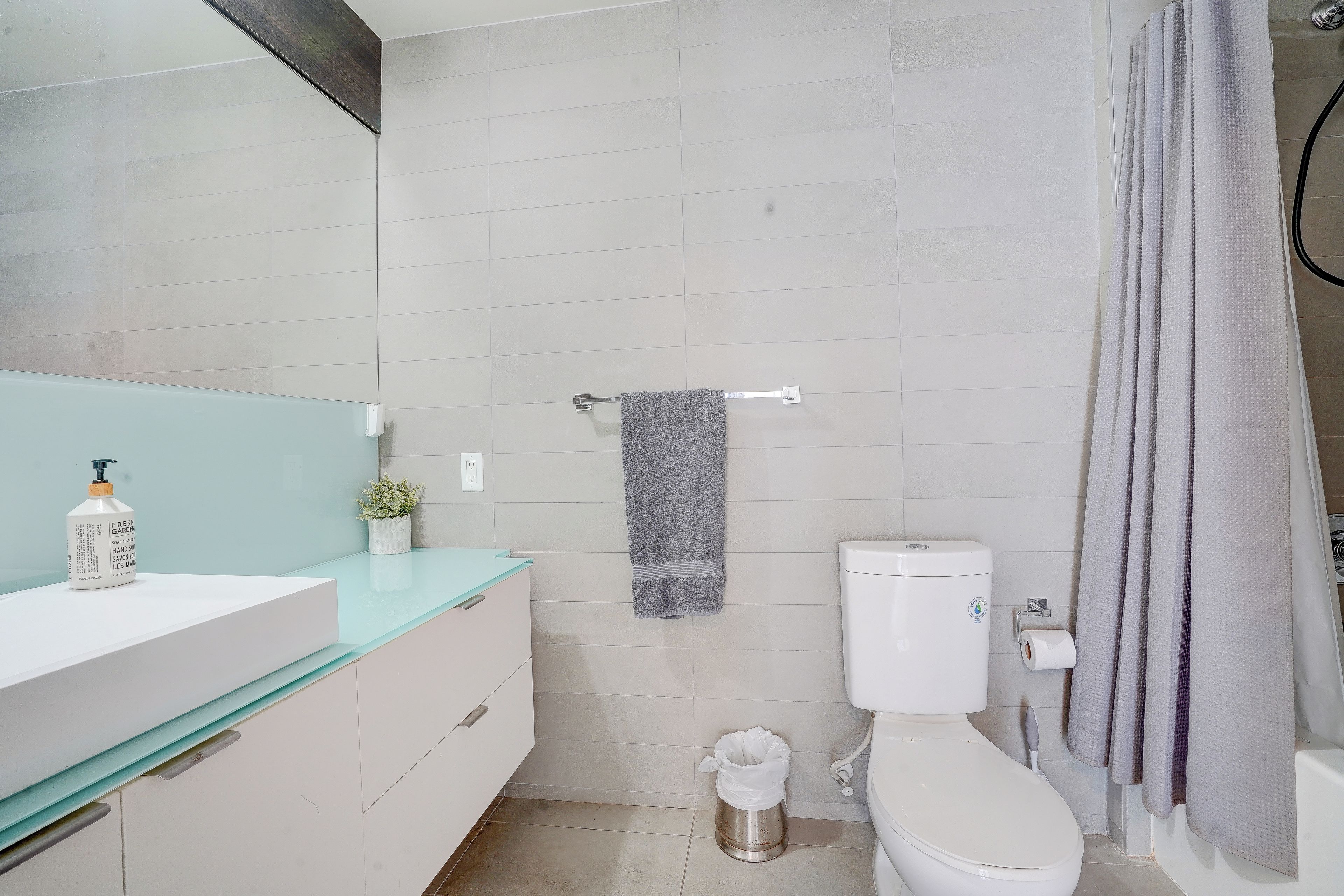 Un baño moderno con inodoro blanco, cabina de ducha con mampara de vidrio y lavamanos blanco con dispensador de jabón.