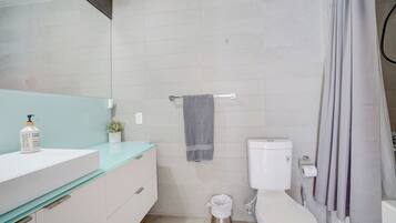 Un baño moderno con inodoro blanco, cabina de ducha con mampara de vidrio y lavamanos blanco con dispensador de jabón.