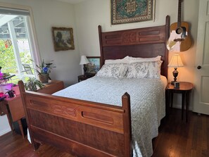 Een slaapkamer met een houten bed, een gitaar, een nachtkastje met een lamp en een raam met planten.