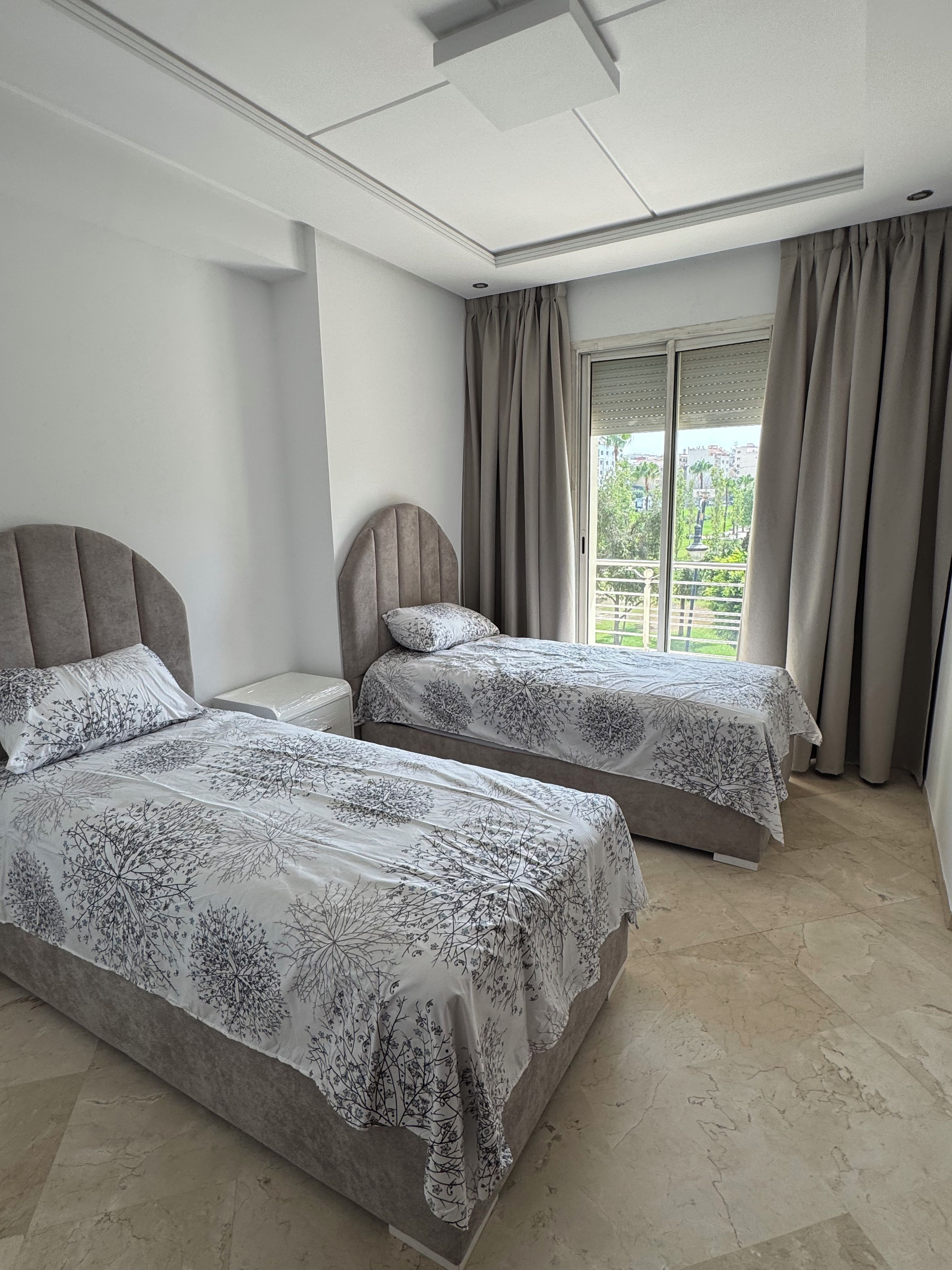 A hotel room with two beds, a large window with curtains, and a view of greenery outside.