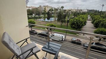 A balcony with two chairs and a small table, overlooking a street with parked cars and a distant view of buildings and a park.