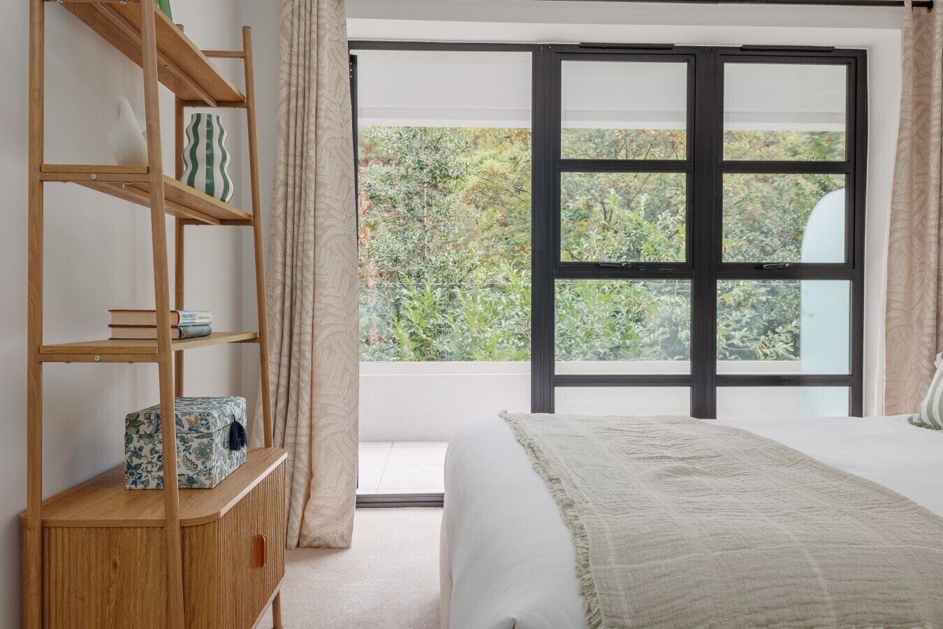 A bedroom with a bed, a wooden bookshelf, and a large window with a view of greenery.