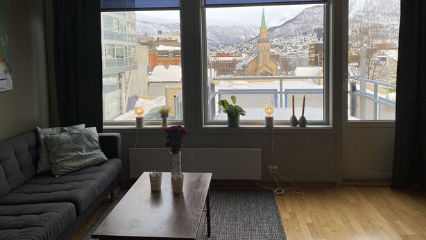 Um quarto com sofá, uma mesa com um vaso, e três janelas com vista para uma cidade e uma igreja.