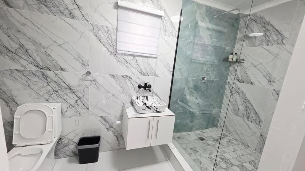 A bathroom with a toilet, a sink with a mirror, a shower with a glass door, and marble walls.
