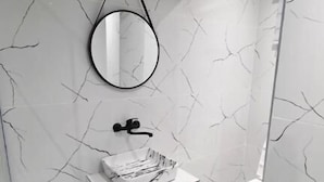 Un baño con paredes de mármol blanco, un espejo redondo, un lavabo blanco y un cubo de basura negro.