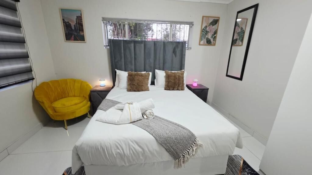 A hotel room with a bed, a yellow armchair, a bedside table with a lamp, and a window with curtains.