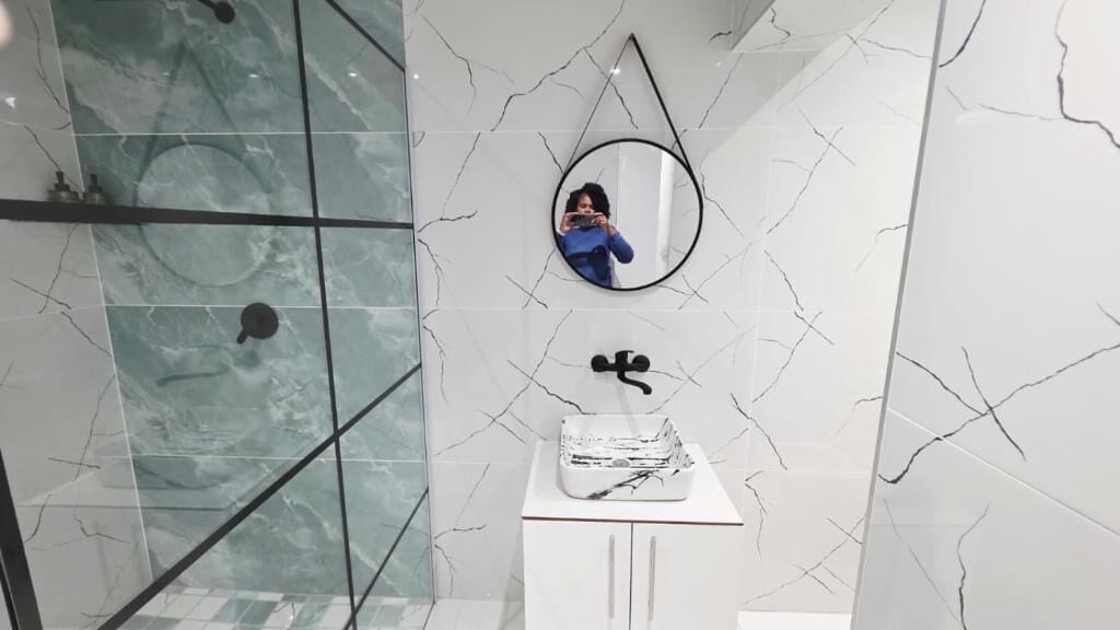 A bathroom with a glass shower enclosure, a white marble wall with a crack pattern, a round mirror, and a white sink with a black faucet.
