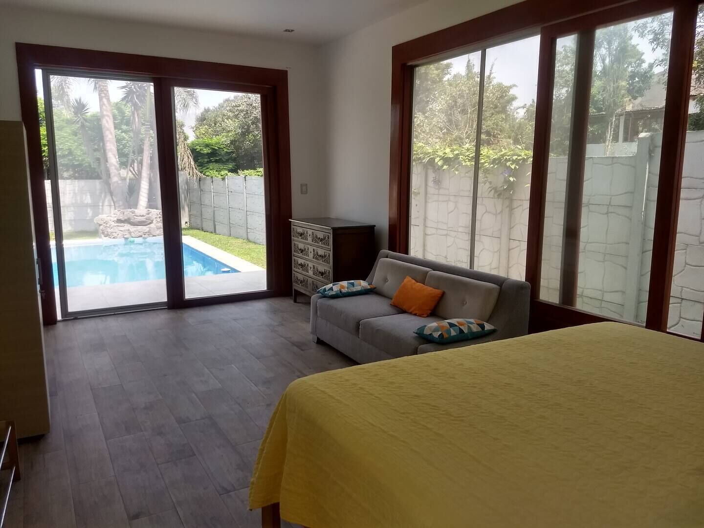 A bedroom with a bed, a sofa, and a view of a swimming pool through a sliding glass door.