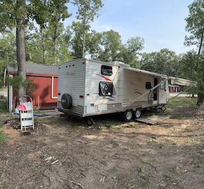 A recreational vehicle parked in a wooded area with a red building nearby. - Lakefront Escape on Twin Lake Cabin + Trailer (Twin Lake)