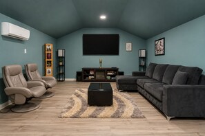 Un salon comprenant un canapé d’angle, deux fauteuils inclinables, un téléviseur à écran plat, une table basse et un climatiseur fixé au mur.