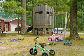 A wooden playhouse with a swing set, a pink toy car, and a green tricycle in a grassy yard with trees. - Lakefront Escape on Twin Lake Cabin + Trailer (Twin Lake)