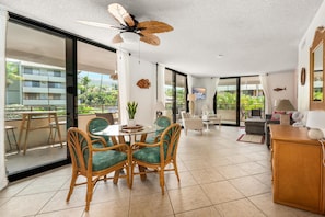 A spacious living area with a dining table surrounded by wicker chairs, a ceiling fan, and a view of a residential building through large glass doors. - Big Island White Sands Village 212 2 Bedroom Condo (Kailua-Kona)