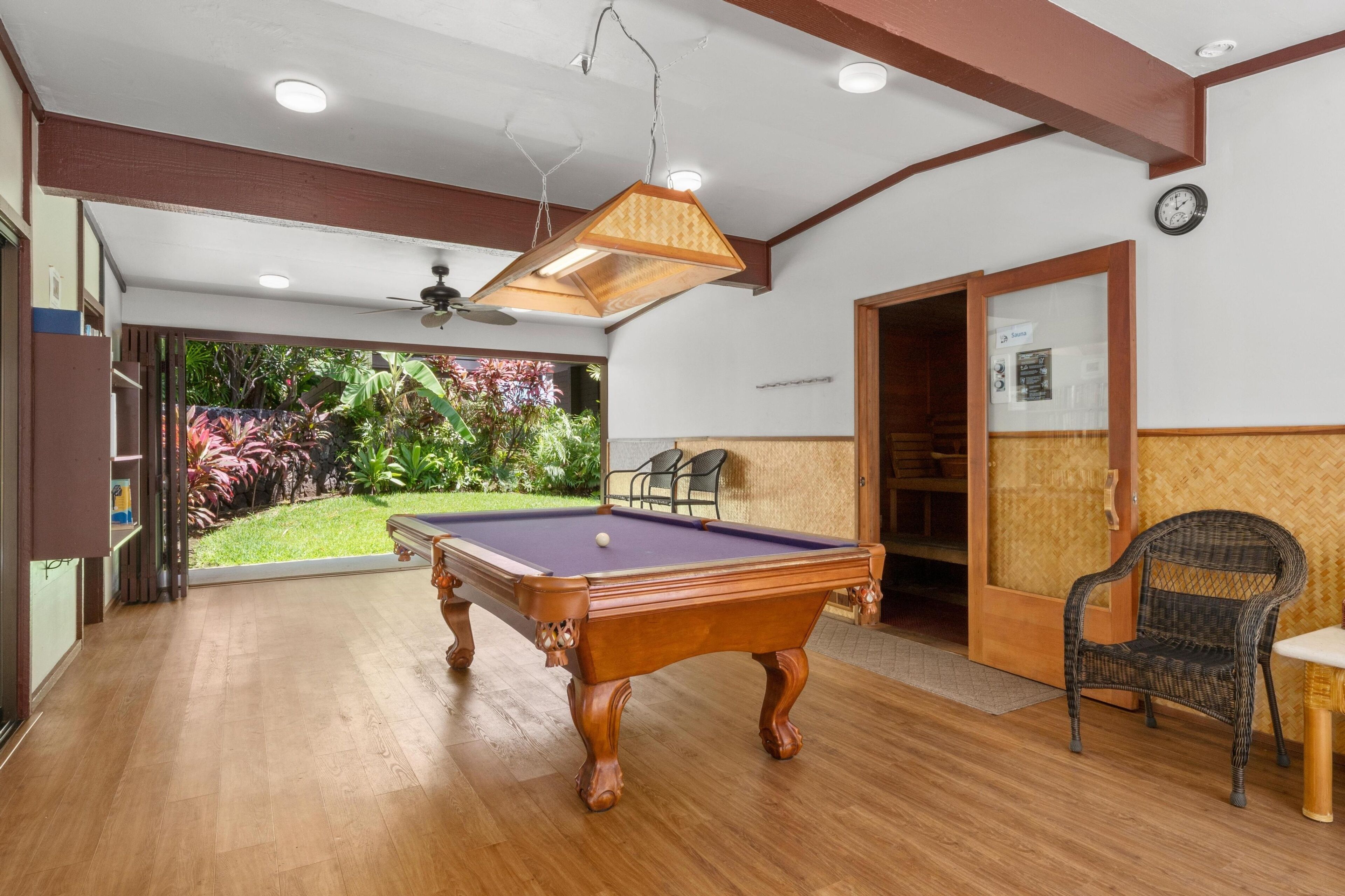 Een pooltafel in een ruimte met houten vloer, een plafondventilator en uitzicht op een tuin.