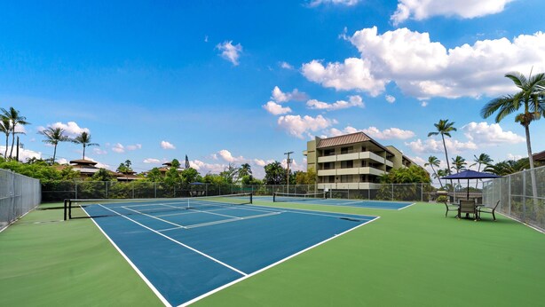 Una cancha de tenis con red, rodeada por una valla, y un edificio al fondo.