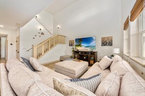 Ein großzügiger Wohnraum mit einem breiten beigefarbenen Sofa, einer Holztreppe und einem an der Wand montierten Flachbildfernseher.