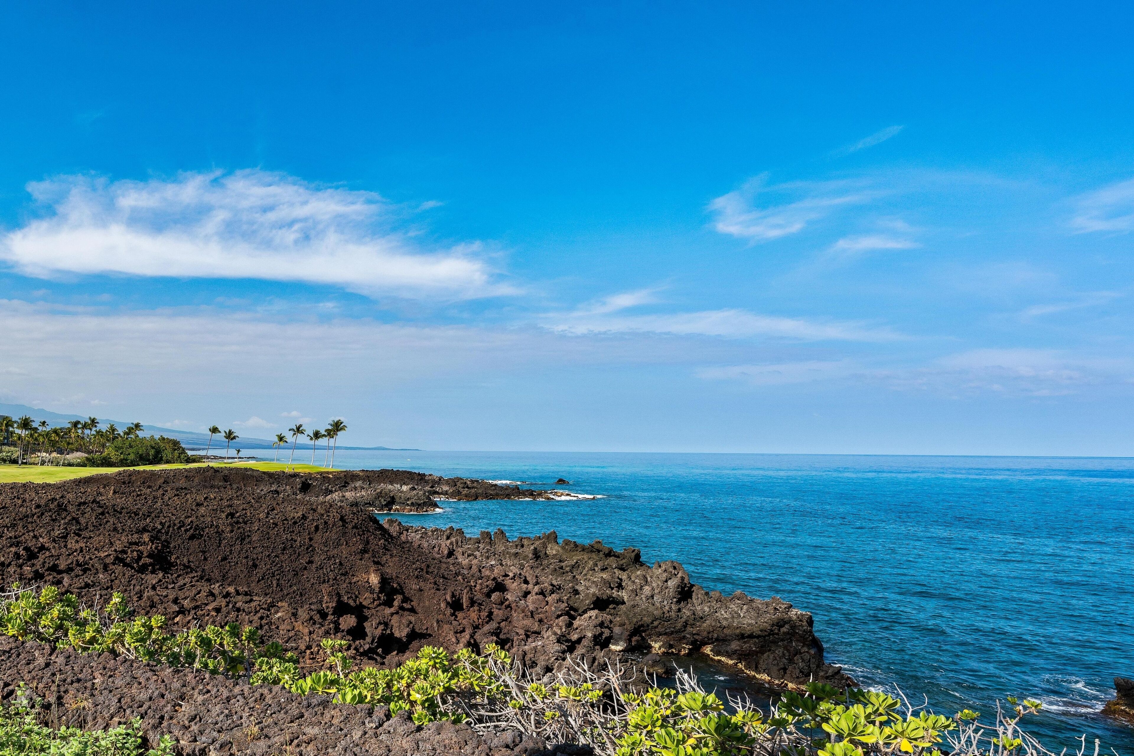 A coastal landscape with a clear sky, a rocky shoreline, and a golf course in the distance.