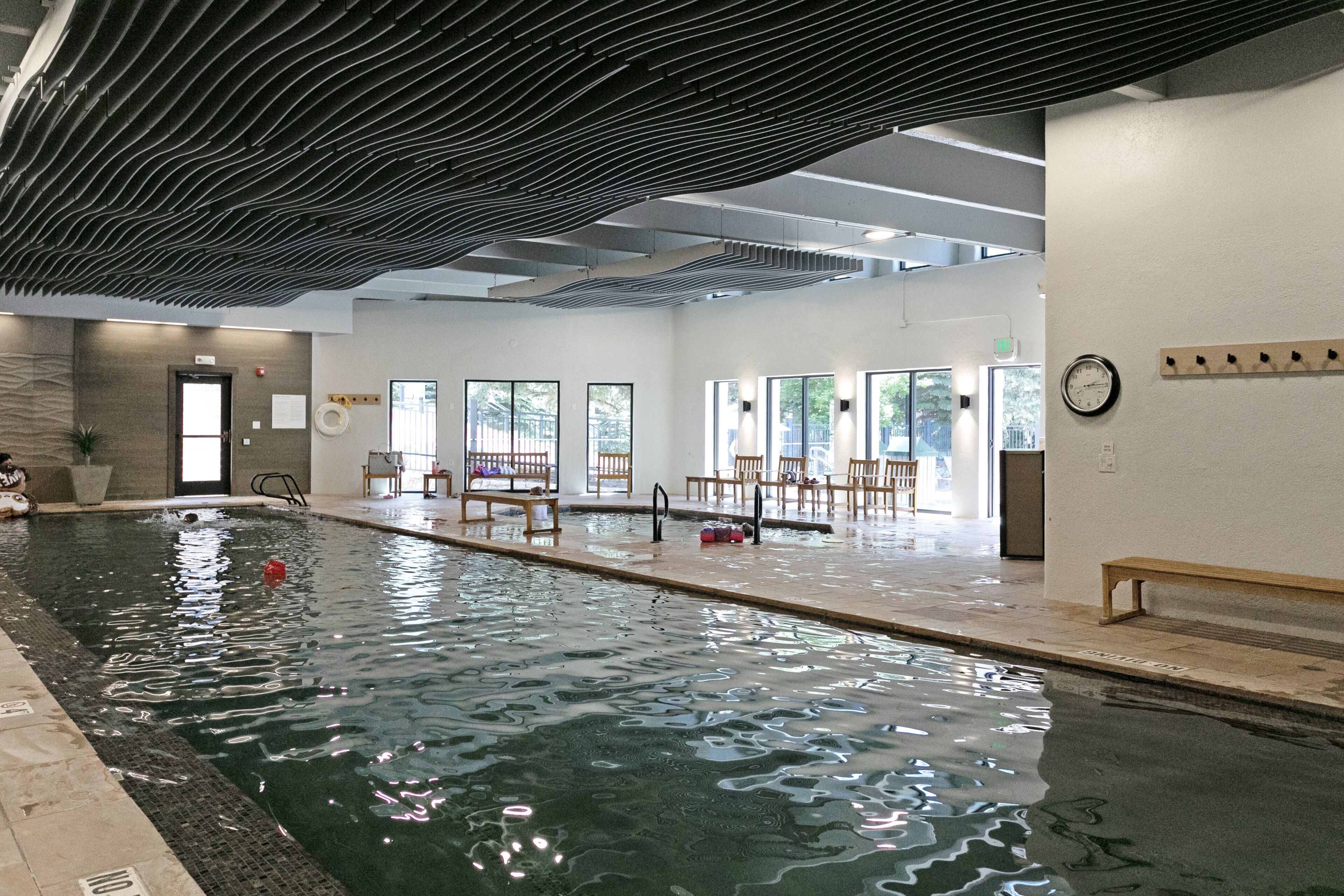 An indoor swimming pool with a bench and chairs, a clock on the wall, and a door leading to another room.
