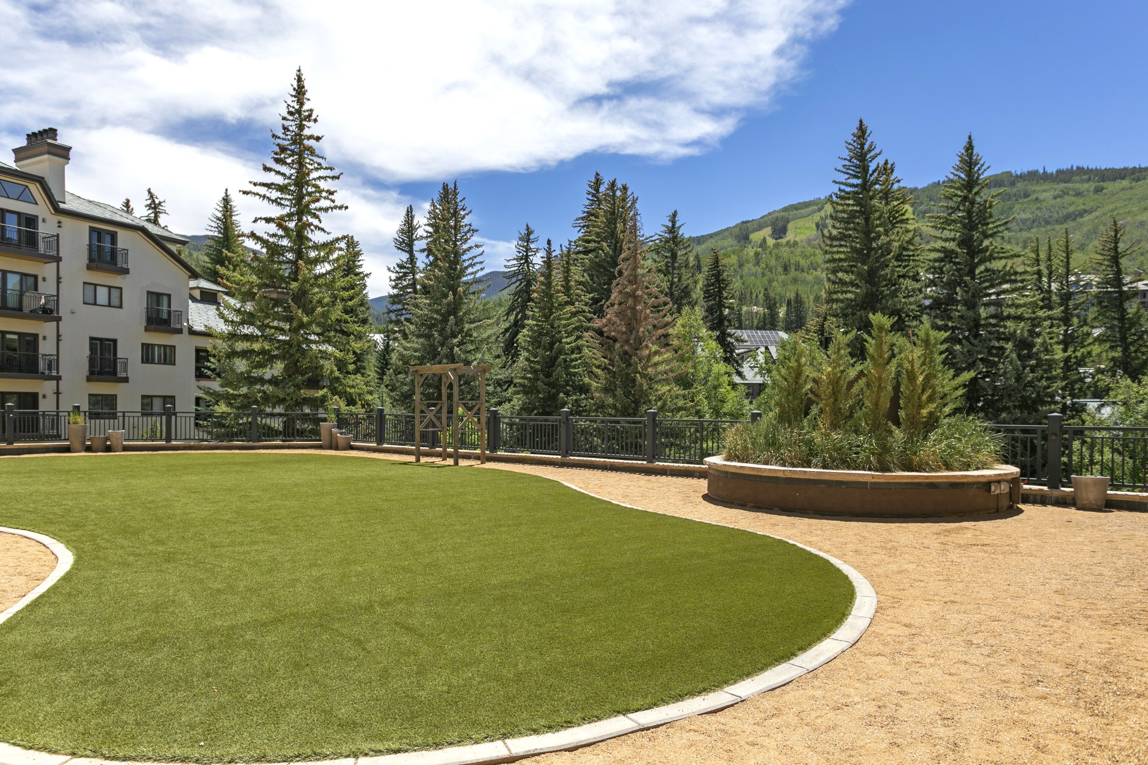 A well-maintained grassy area with a wooden pergola, surrounded by a metal fence and tall evergreen trees.