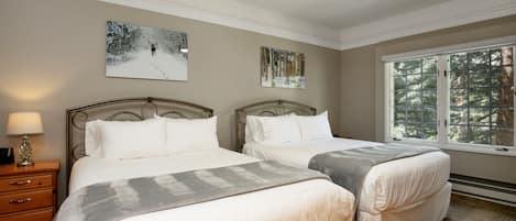 A bedroom with two beds, a nightstand with a lamp, and a window with a view of trees.