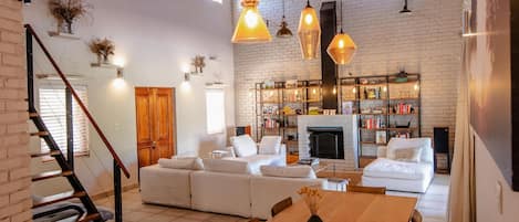 A spacious living area with a wooden ceiling, white brick walls, and a fireplace. There is a dining table with chairs and a staircase leading to another level.