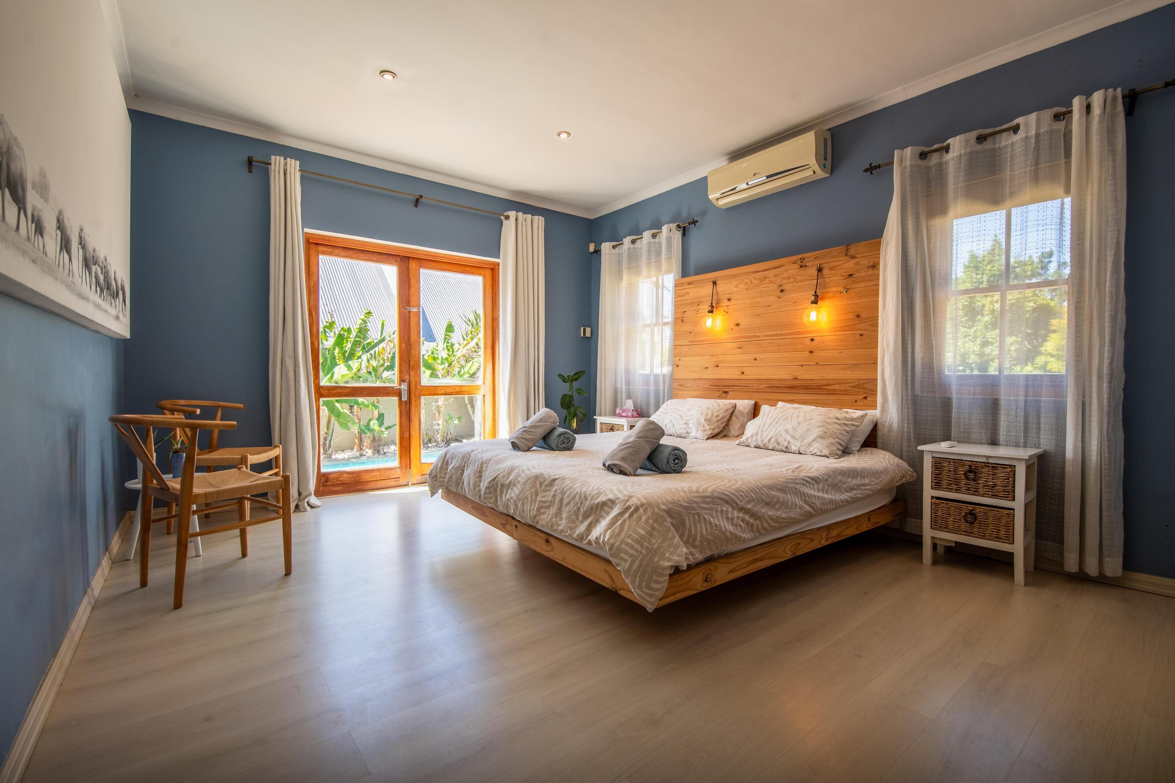 Een slaapkamer met een houten bed, een bureau en een stoel, een groot raam met gordijnen, en uitzicht op groen buiten.