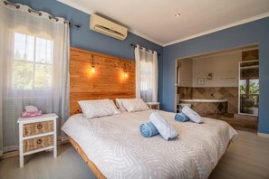 Een slaapkamer met een houten hoofdeinde, een bed met beddengoed met patroon, een nachtkastje, een wandairconditioner en een badkamer met een bad en een raam.