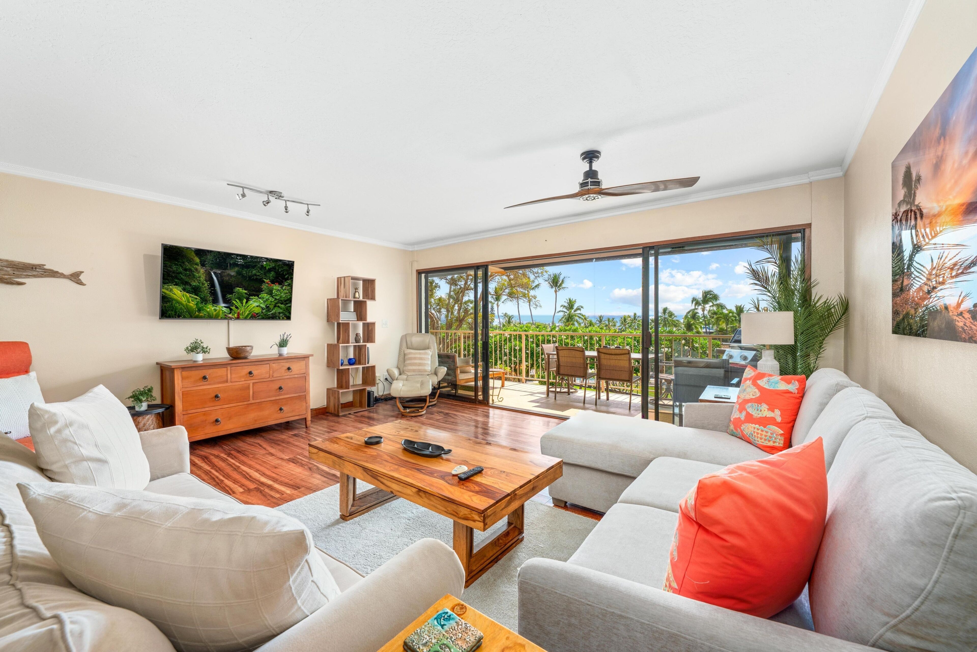 A spacious living room with a large TV, wooden furniture, a ceiling fan, and a view of the outdoors through a sliding glass door.