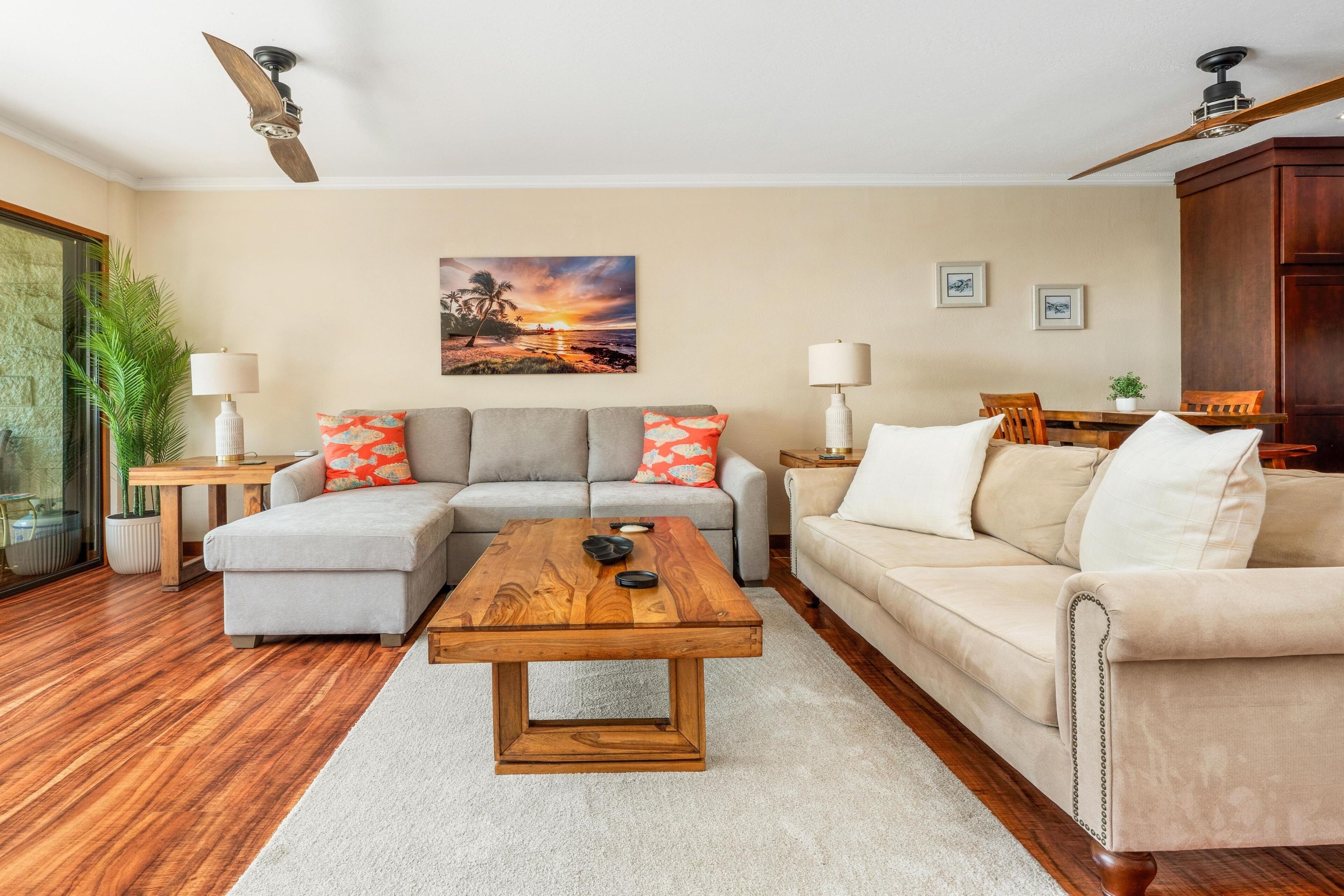 Un salon avec une table basse en bois, deux canapés, un tableau représentant un coucher de soleil et un ventilateur de plafond.