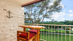 A wooden table and chair set on a balcony with a stone wall and a view of a well-maintained garden and trees. - Big Island Keauhou Punahele C202 2 Bedroom Condo (Kailua-Kona)