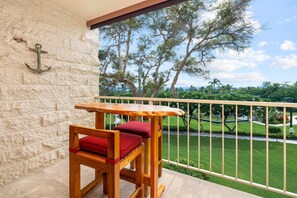 A wooden table and chair set on a balcony with a stone wall and a view of a well-maintained garden and trees. - Big Island Keauhou Punahele C202 2 Bedroom Condo (Kailua-Kona)