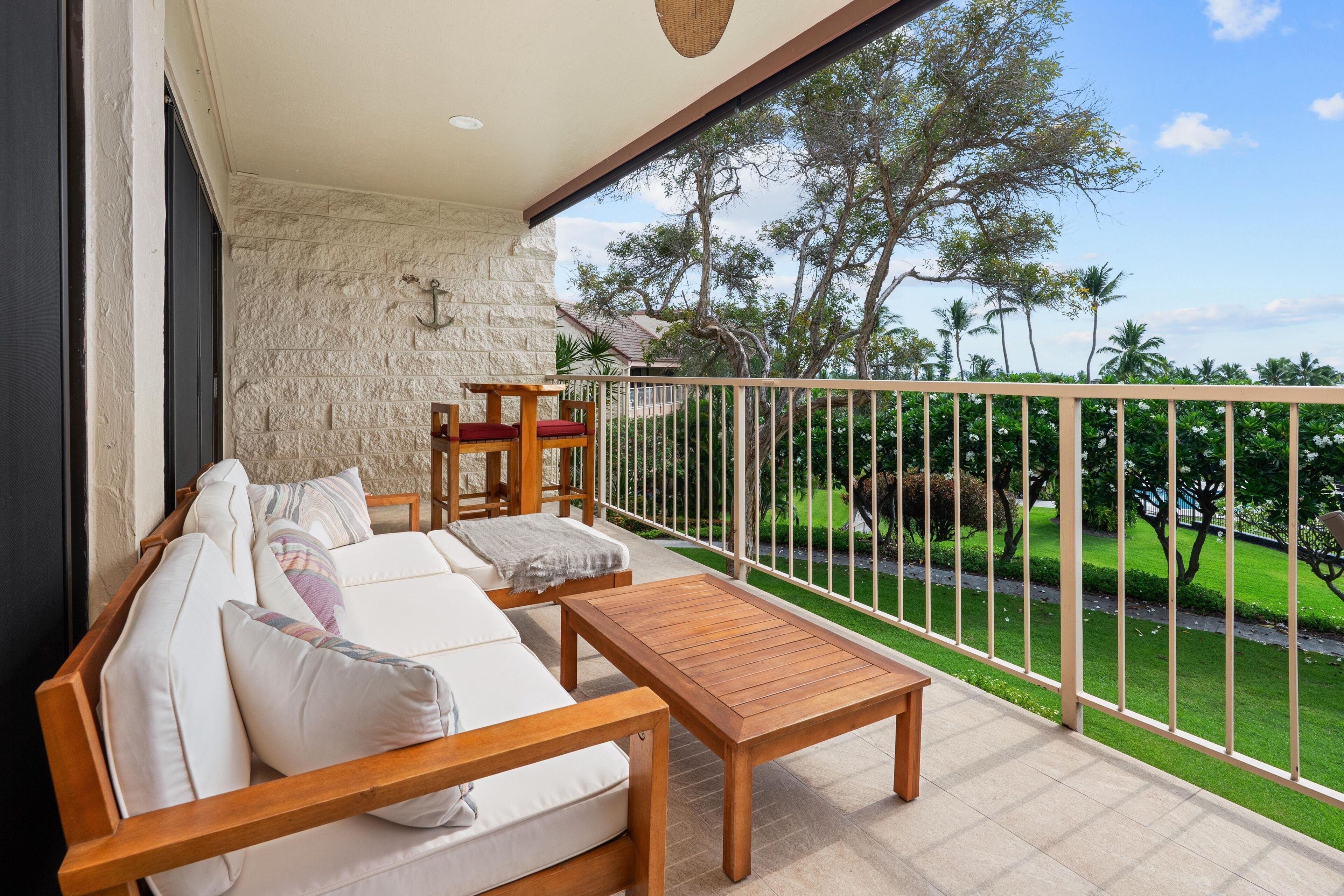 A balcony with a wooden sofa, a small table, and a view of a well-maintained garden and palm trees.