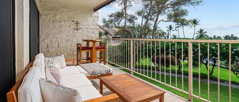 A balcony with a wooden sofa, a small table, and a view of a well-maintained garden and palm trees.
