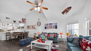 Un salon moderne doté d’un ventilateur de plafond, un coin repas avec une table et des chaises blanches, et une cuisine avec des meubles blancs.