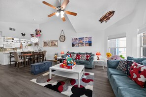A modern living room with a ceiling fan, a dining area with a white table and chairs, and a kitchen with white cabinets. - your Happy Place Close To All The Magic! 3 Bedroom Condo (Kissimmee)