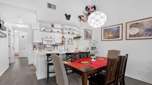 Une cuisine moderne avec des meubles blancs, un coin repas doté d’une table rouge et des éléments de décoration murale, notamment des oreilles de Mickey Mouse et une horloge.