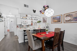 A modern kitchen with white cabinetry, a dining area with a red table, and wall decorations including Mickey Mouse ears and a clock. - your Happy Place Close To All The Magic! 3 Bedroom Condo (Kissimmee)