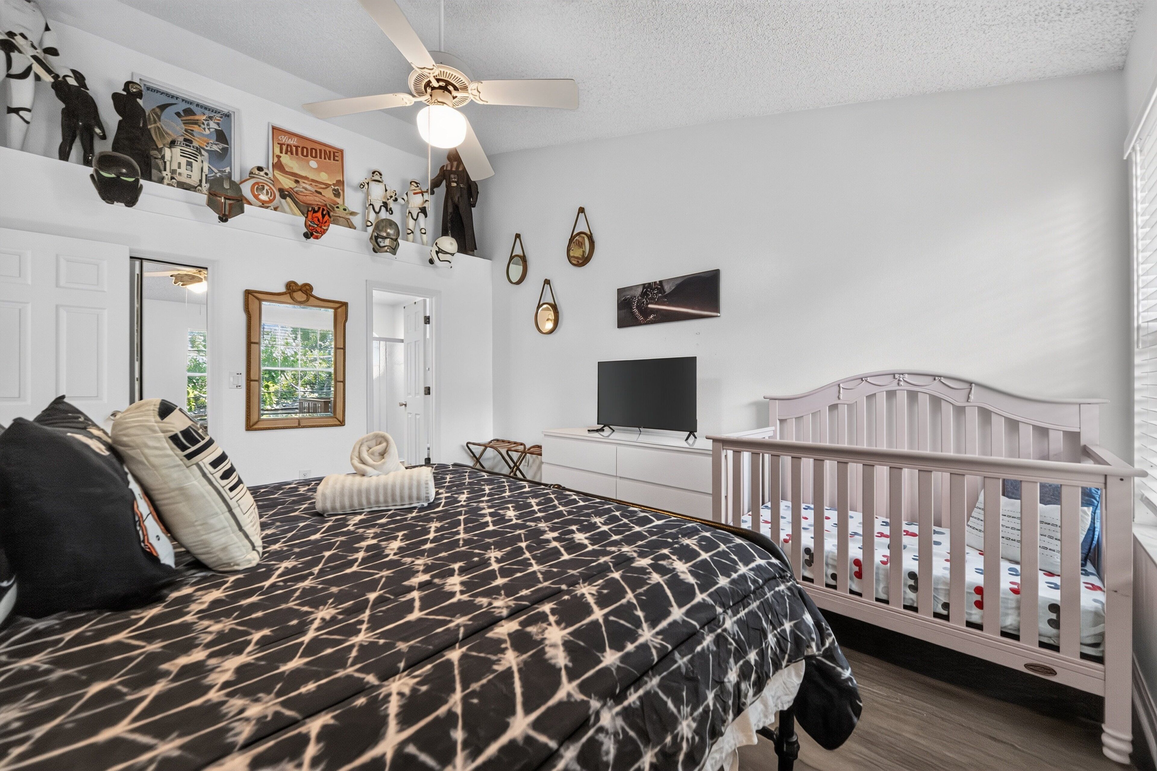 A bedroom with a bed, a crib, a ceiling fan, wall decorations, and a television.