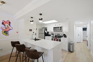 A modern kitchen with a central island, bar stools, a refrigerator, microwave, and a trash can. - "Castle Retreat" Close To All The Magic! 3 Bedroom Condo (Kissimmee)