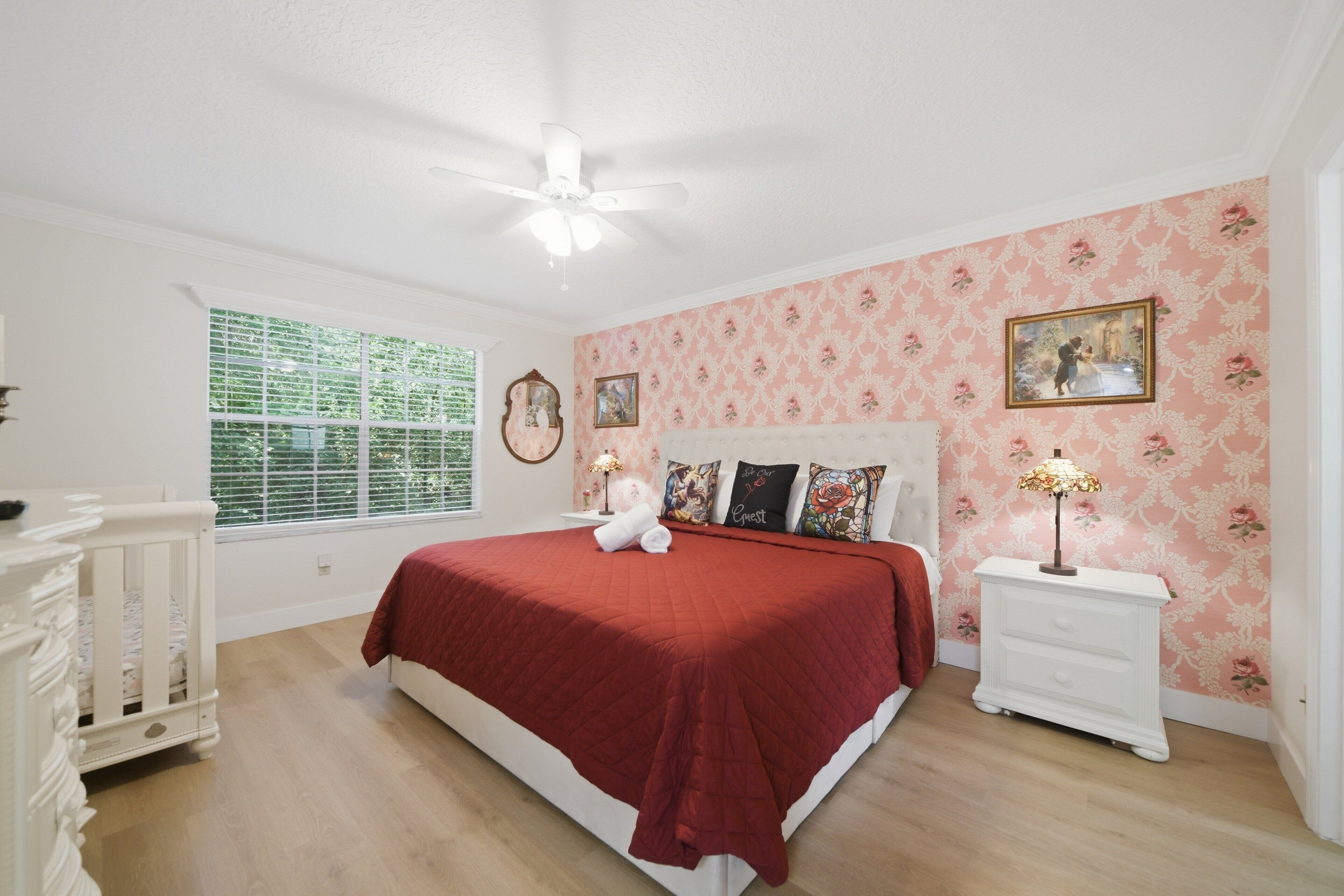 A bedroom with a large bed, a nightstand, a dresser, a window with blinds, and floral wallpaper.