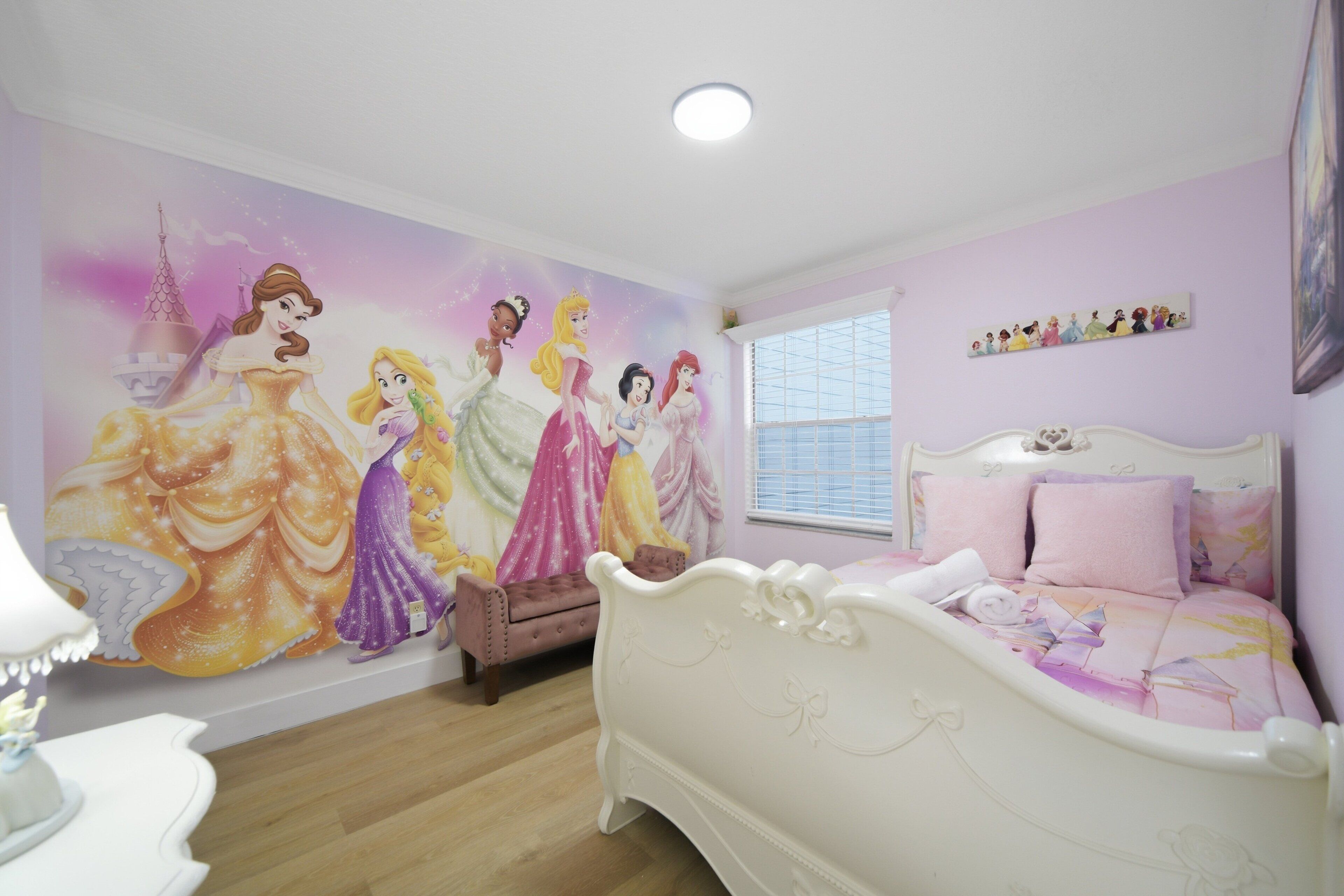 A bedroom with a large wall mural of Disney princesses, a bed with pink bedding, and a window with blinds.