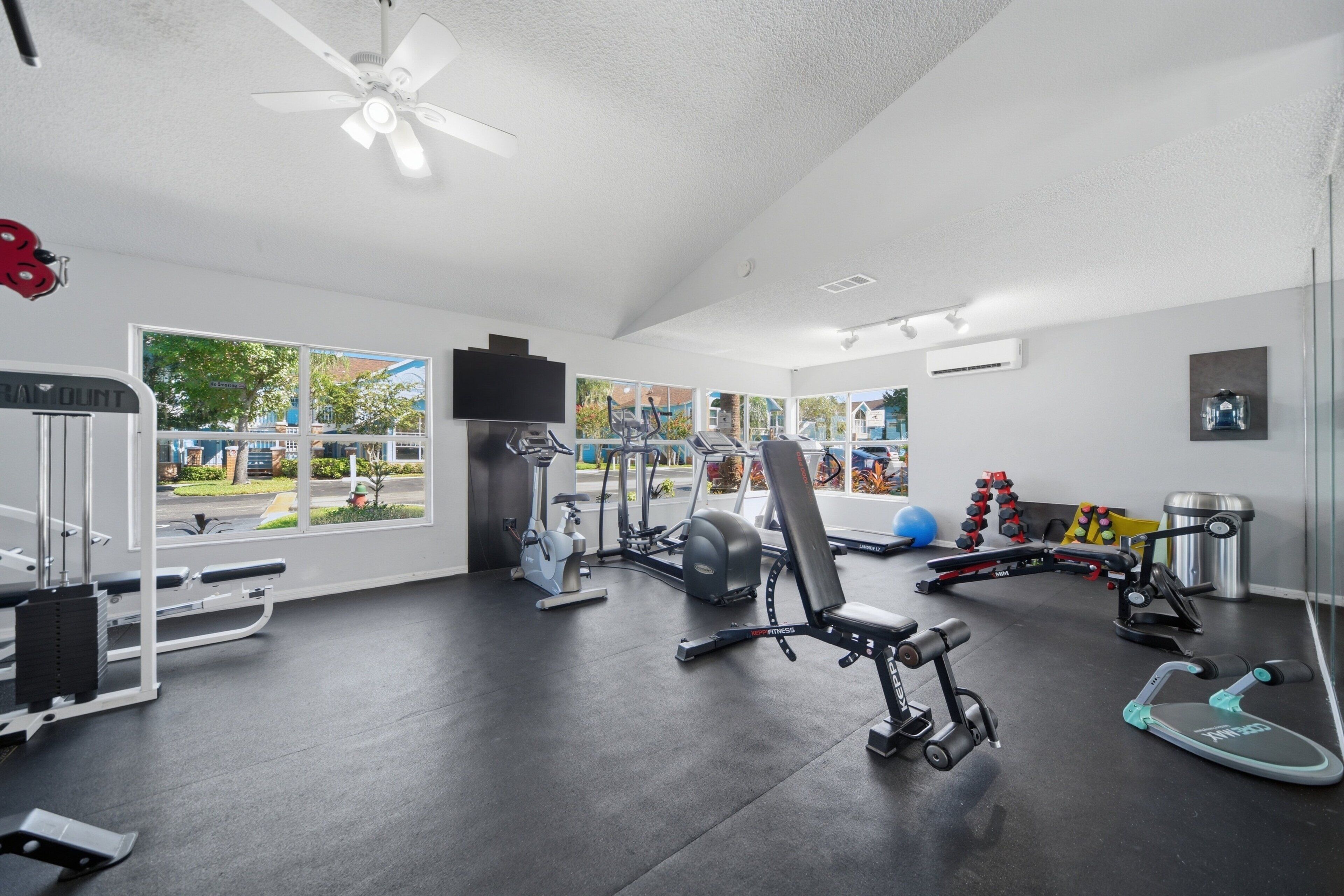 Ett gym med olika typer av träningsutrustning, en takfläkt och stora fönster som ger utsikt över omgivningen.